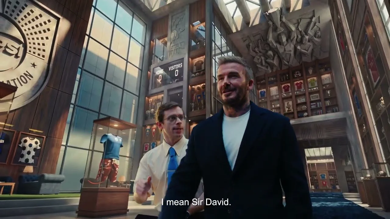 David Beckham caminha e sorri para a frente, em um grande museu de futebol e hospitalidade, com uma pessoa mais jovem ao seu lado.