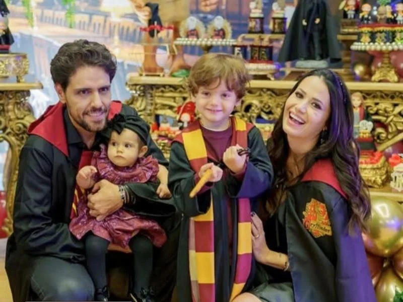 Foto de estúdio de uma família de quatro pessoas em trajes de Harry Potter. O pai e a mãe usam mantos pretos e vermelhos, o filho mais velho um cachecol Grifinória e a bebê um vestido roxo. Todos sorriem em frente a um fundo temático com prateleiras e decorações.