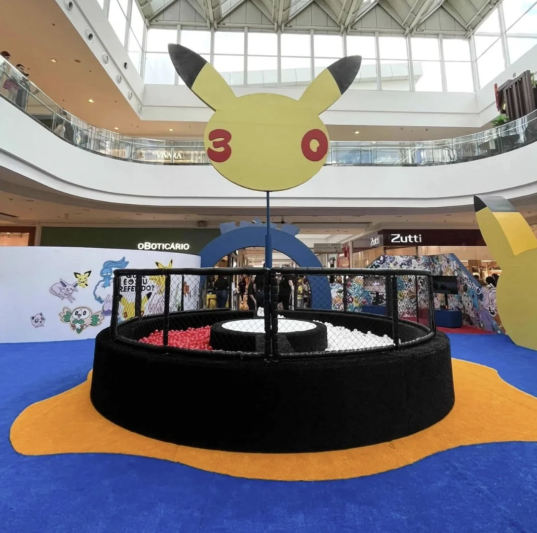 Vista frontal de uma atração de shopping com o tema Pokémon. O elemento principal é uma grande escultura suspensa da cabeça do Pikachu com bochechas vermelhas circulares com os números "3" e "0". Abaixo, uma piscina de bolinhas circular com divisões vermelhas e brancas, simulando uma Pokébola, é cercada por uma grade preta e posicionada sobre um carpete azul com um tapete amarelo recortado. O fundo mostra outras decorações do Pokémon e lojas do shopping.