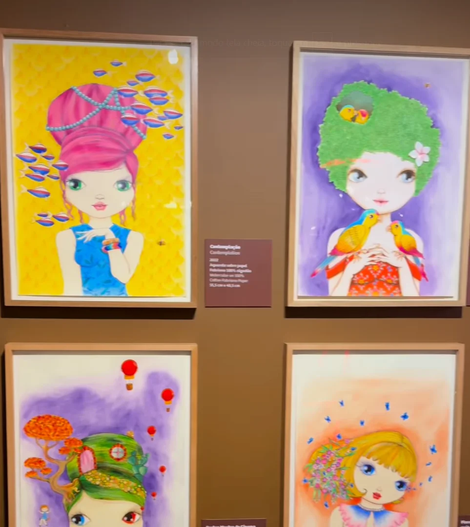 Quatro retratos ilustrados de mulheres, emoldurados e dispostos em duas fileiras, estão pendurados em uma parede marrom em uma galeria. Cada obra apresenta uma paleta de cores diferente e elementos distintos em seus cabelos e roupas.