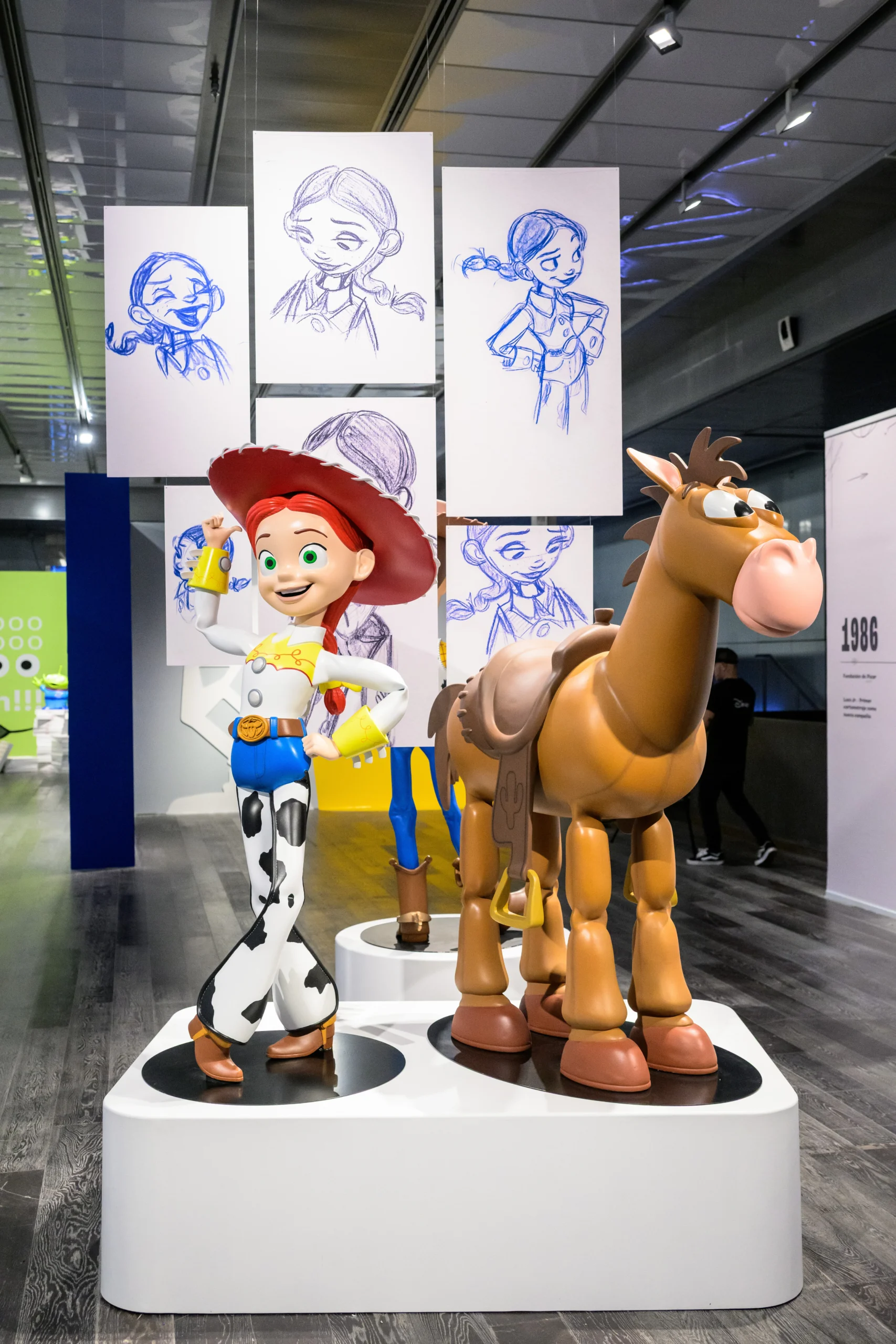Uma estátua em tamanho real de Jessie e Bala no Alvo, de Toy Story, em exibição em uma exposição da Pixar. A estátua de Jessie está posando com um braço levantado e um pé sobre o plinto, enquanto Bala no Alvo está ao lado dela. Vários desenhos de esboço de Jessie estão pendurados acima.