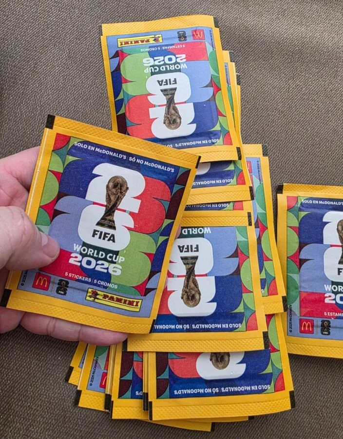 Mão segurando um pacotinho de figurinhas da Copa do Mundo FIFA 2026 da Panini, com outros pacotes empilhados ao fundo. O envelope é amarelo com detalhes coloridos e o logotipo oficial do torneio.