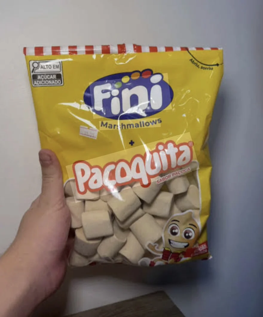 Uma mão segura um pacote amarelo de Marshmallows Fini sabor Paçoquita contra uma parede branca. O pacote está levemente amassado e mostra os marshmallows beges através da transparência frontal.