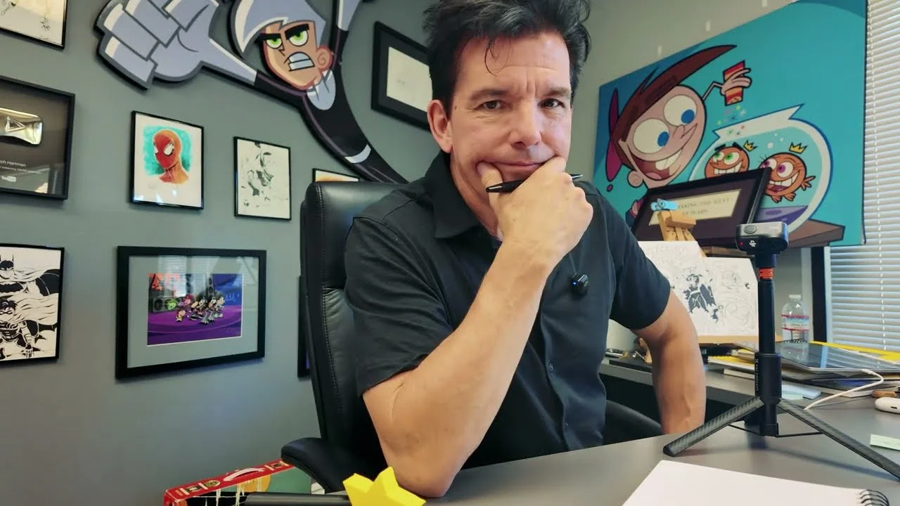 Butch Hartman, criador de desenhos animados, sentado em sua mesa de trabalho cercado por artes de suas criações, como Danny Phantom e Os Padrinhos Mágicos.