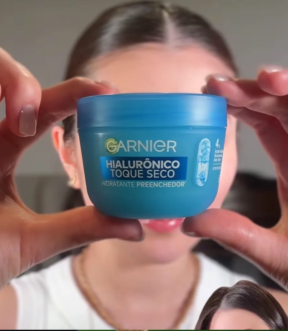 Uma pessoa segura um pote de creme facial azul da marca Garnier, com o rótulo 'Hialurônico Toque Seco'. O produto está centralizado em frente ao rosto da pessoa, que serve de fundo desfocado.