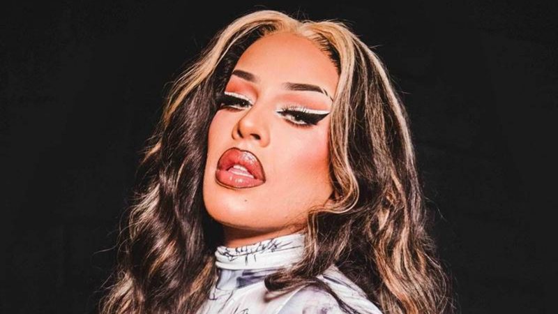 Retrato de busto da Gloria Groove contra um fundo escuro. Ela posa com a cabeça levemente inclinada, exibindo cabelos longos ondulados com mechas loiras frontais, lábios contornados e um delineado branco duplo sobre a pálpebra.