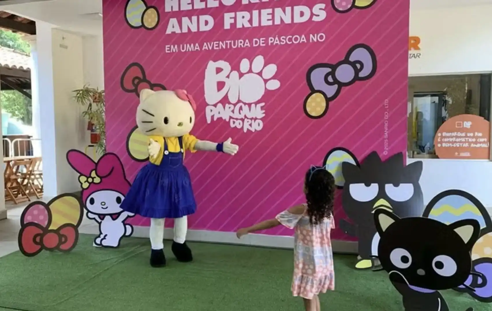 Uma criança de vestido de bolinhas encontra uma pessoa fantasiada de Hello Kitty em frente a um painel rosa da BioParque do Rio.