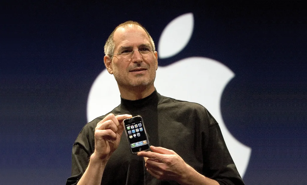 Uma foto de Steve Jobs no palco, vestindo seu característico casaco de gola alta preto, segurando um iPhone original em sua mão direita. Ele está sorrindo levemente e olhando para fora da câmera, com um grande logotipo branco da Apple em um fundo azul escuro atrás dele.