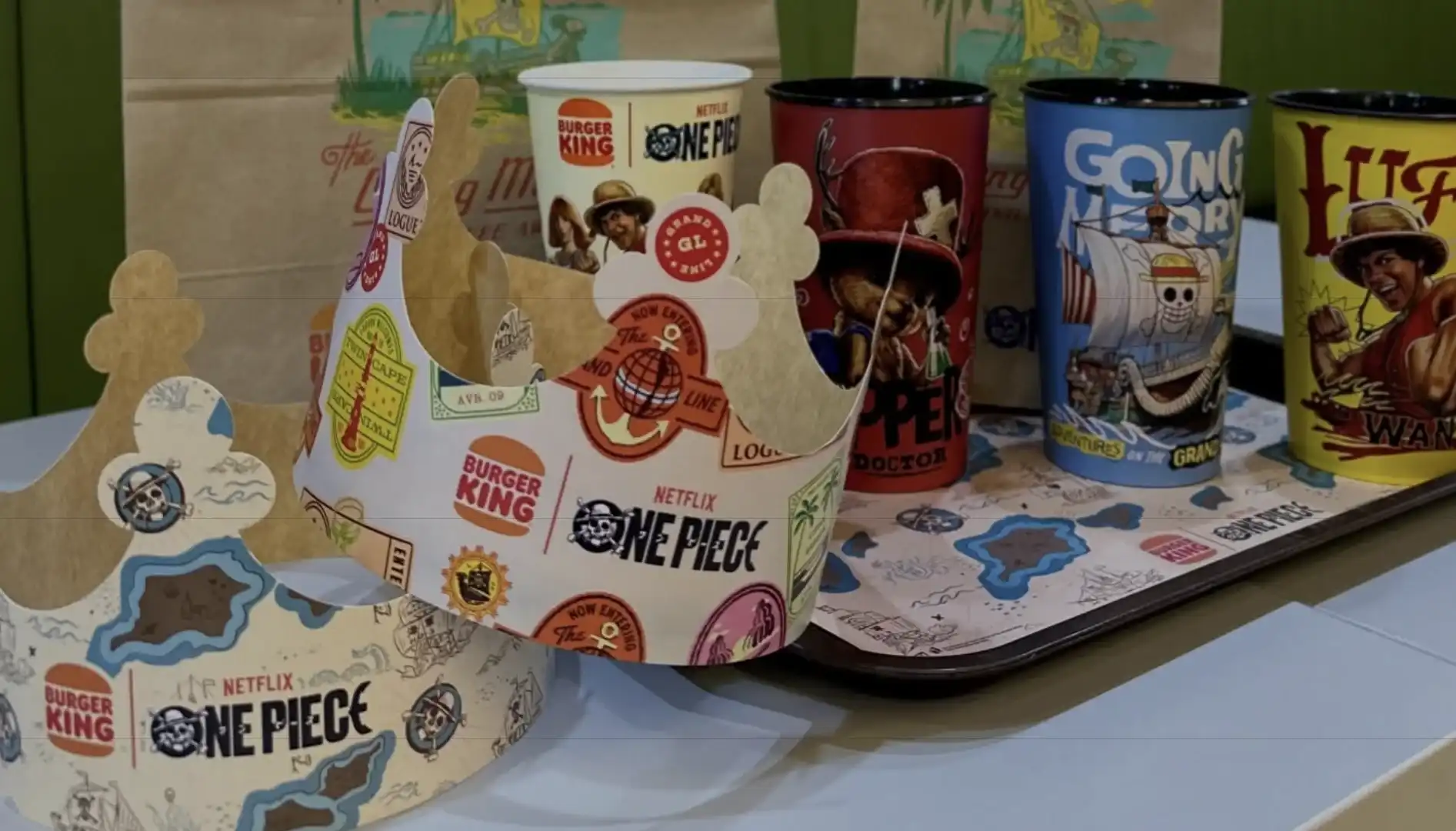 Uma foto de perto, tirada do nível dos olhos, mostra dois coroas de papelão do Burger King e três copos, todos inspirados em "One Piece", em cima de um prato. O prato está em cima de uma mesa verde.