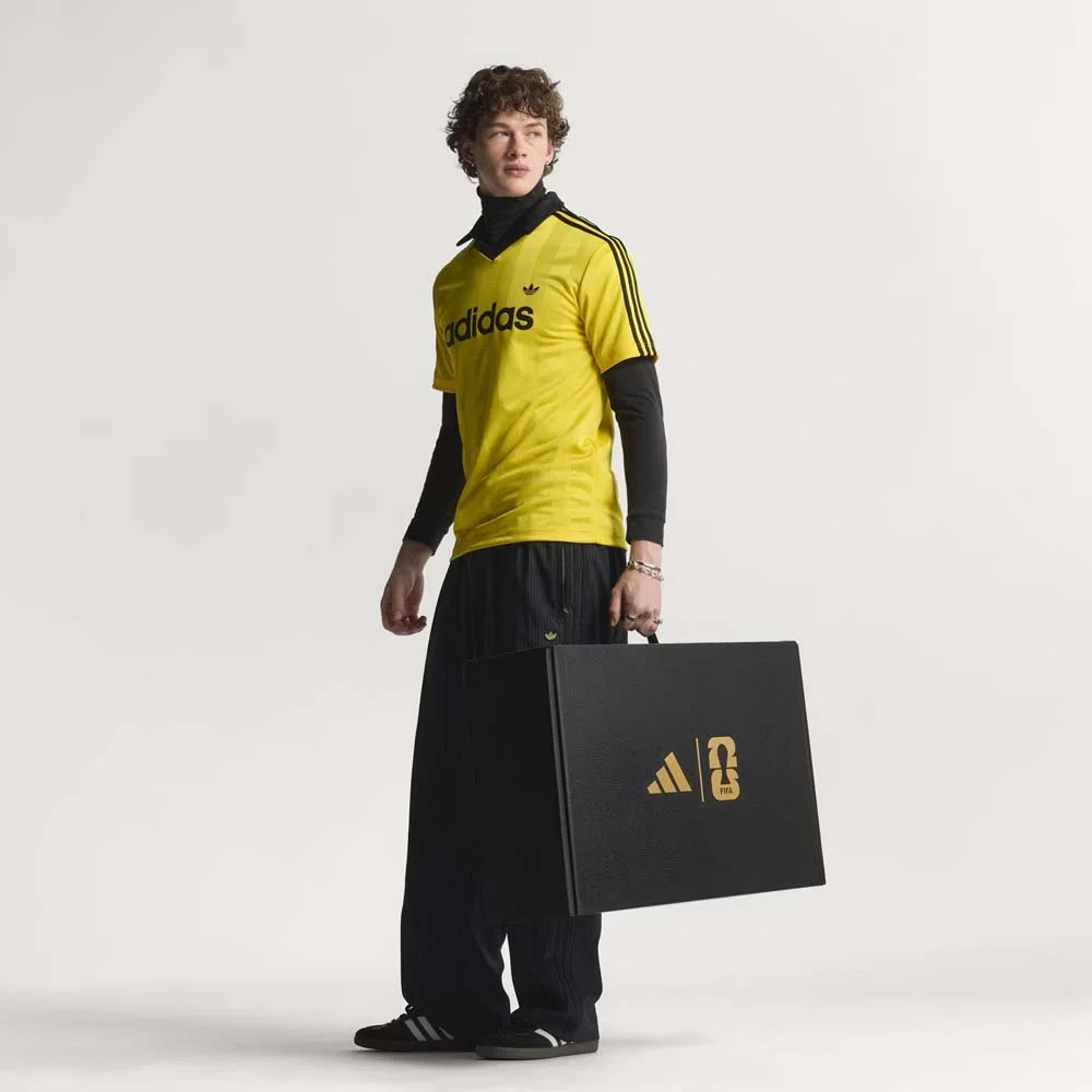 Um modelo masculino em pé com cabelos cacheados, olhando para a esquerda, segurando uma grande caixa preta na mão esquerda contra um fundo cinza claro. Ele veste uma camisa polo amarela brilhante da Adidas com três listras pretas nas mangas e o logotipo da Adidas no peito, sobre uma blusa de gola alta preta de manga longa, calças de moletom pretas largas e tênis pretos. A caixa preta que ele segura tem os logotipos dourados da Adidas e da FIFA no centro.