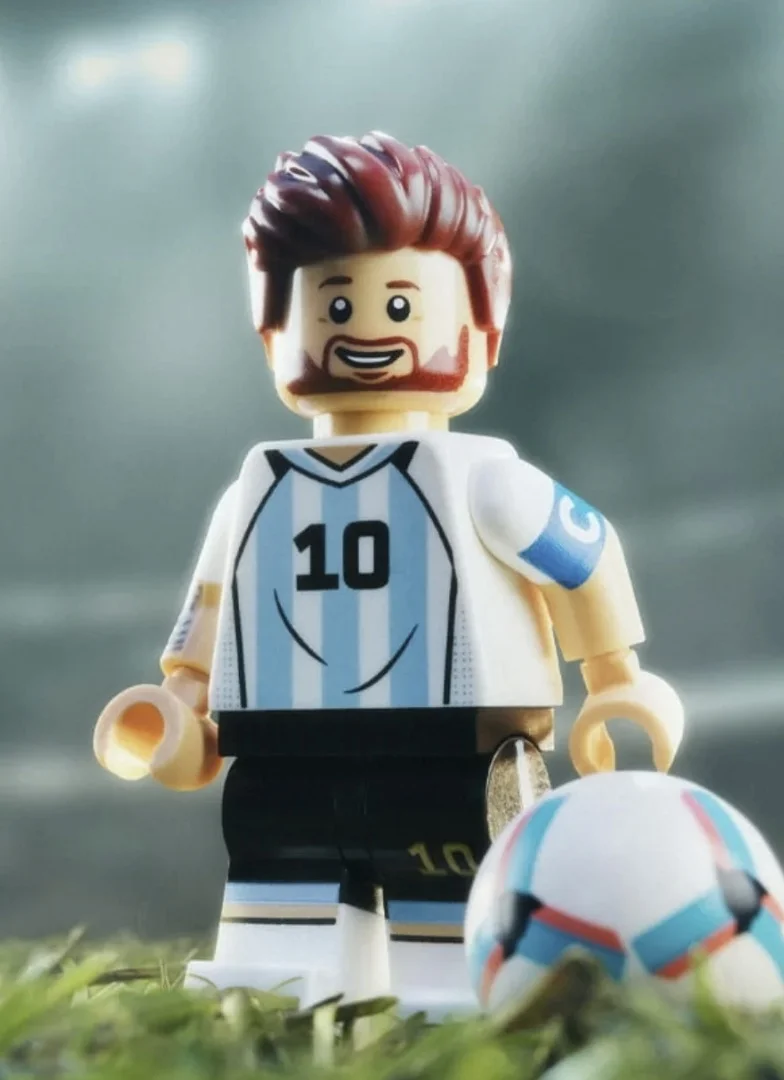 Uma foto em close-up de uma minifigura LEGO estilizada de Lionel Messi vestindo o kit da seleção da Argentina, de pé sobre um campo de futebol de grama.