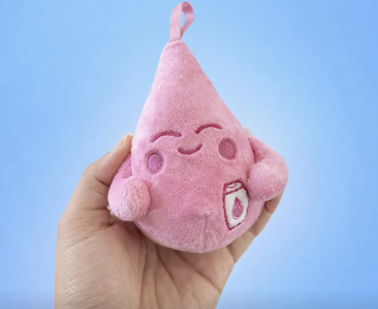 Uma mão segura uma pequena pelúcia rosa em formato de gota. O boneco tem um rosto sorridente bordado, bochechas rosadas e um pequeno detalhe de frasco de xampu no corpo. O fundo é azul claro e sólido.