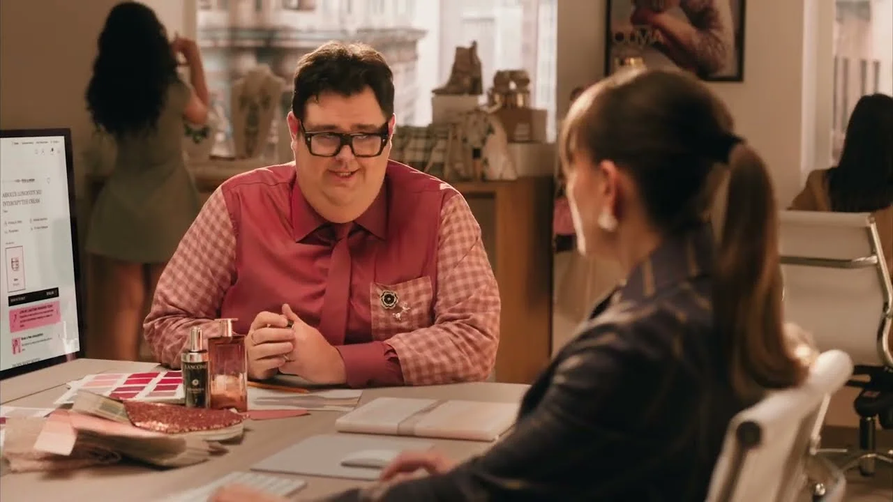 Um homem de óculos e camisa rosa xadrez está sentado a uma mesa, conversando com uma mulher de costas com rabo de cavalo. A mesa tem cosméticos e tecidos. No fundo, há mais pessoas e um computador.