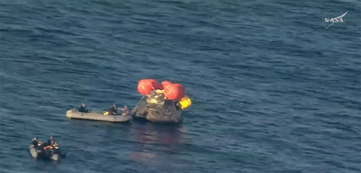 Uma imagem de satélite de ângulo baixo de uma equipe de recuperação da Marinha dos EUA aproximando-se da espaçonave Orion flutuando na vertical no Oceano Pacífico em dois barcos infláveis de casco rígido (RHIBs).