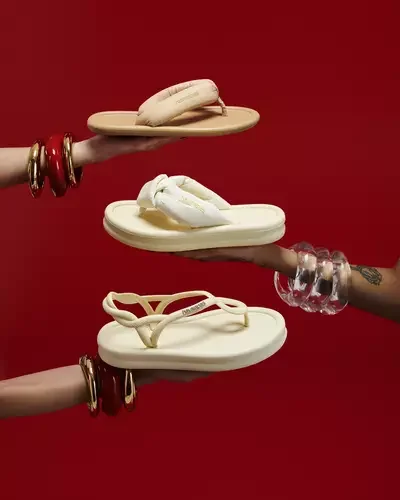 Uma composição fotográfica de estúdio com três mãos femininas, cada uma estendida e segurando um estilo diferente de sandália Havaianas de cor creme. As mãos estão dispostas verticalmente contra um fundo vermelho vivo. A mão superior usa pulseiras de ouro e resina vermelha, a do meio usa pulseiras de plástico transparente e a inferior usa pulseiras de metal e resina. As sandálias são: uma rasteirinha de dedo simples acolchoada (superior), uma sandália plataforma de dedo com nó acolchoado (meio) e uma sandália rasteirinha de tiras trançadas (inferior).