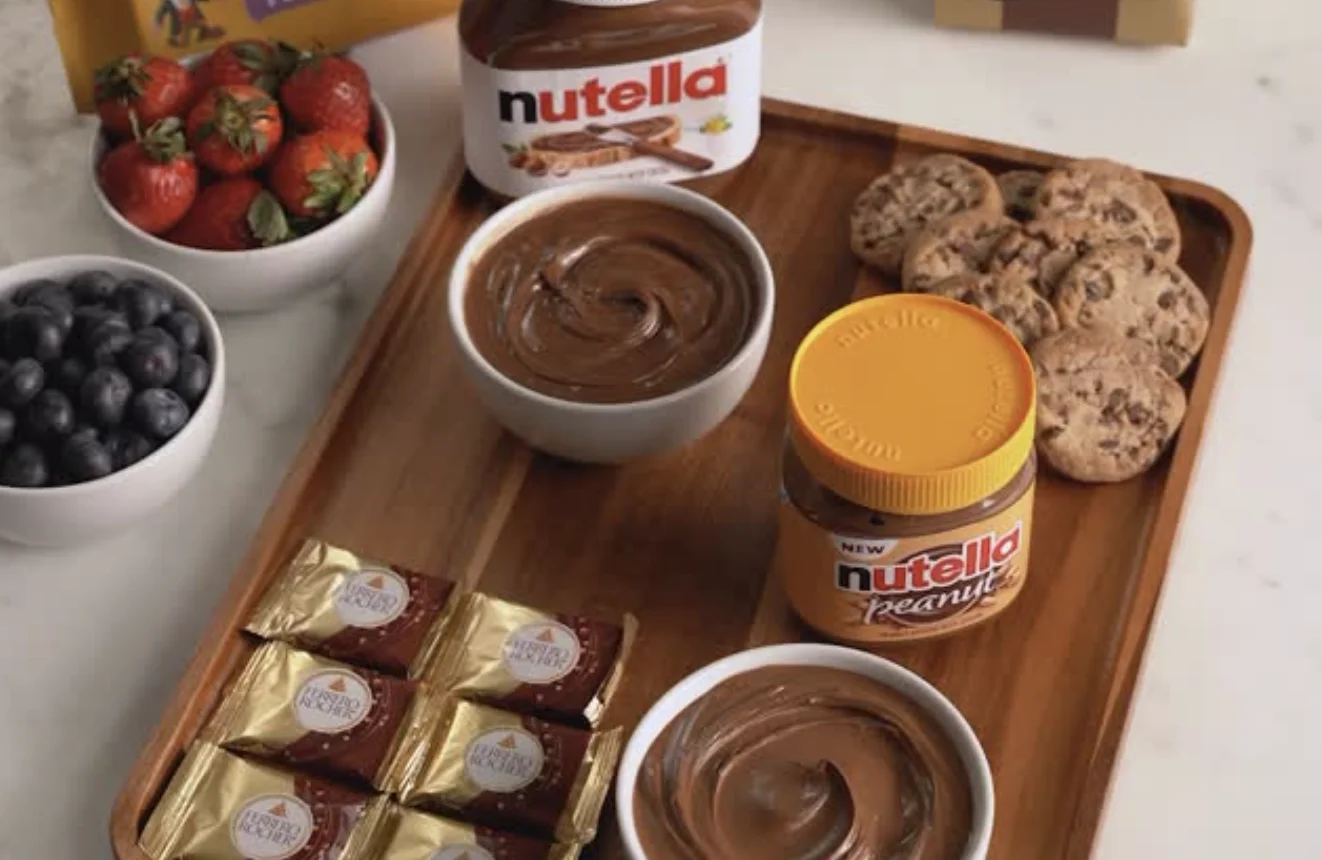 Uma tábua de madeira com potes de Nutella original e Nutella Peanut, acompanhados de morangos, mirtilos, cookies e chocolates Ferrero Rocher.