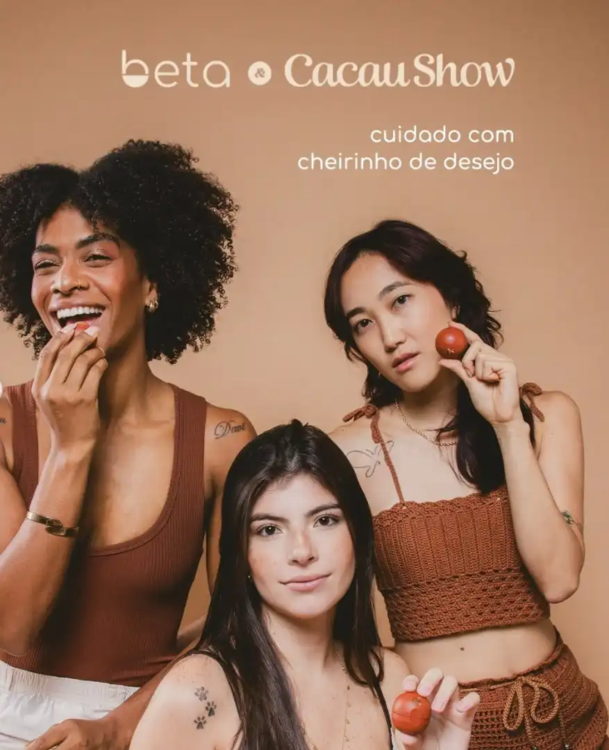Três mulheres de diferentes etnias posando juntas em frente a um fundo bege claro. Duas seguram a esfera do hidratante labial beta Cacau Show. O logotipo da colaboração está no topo.