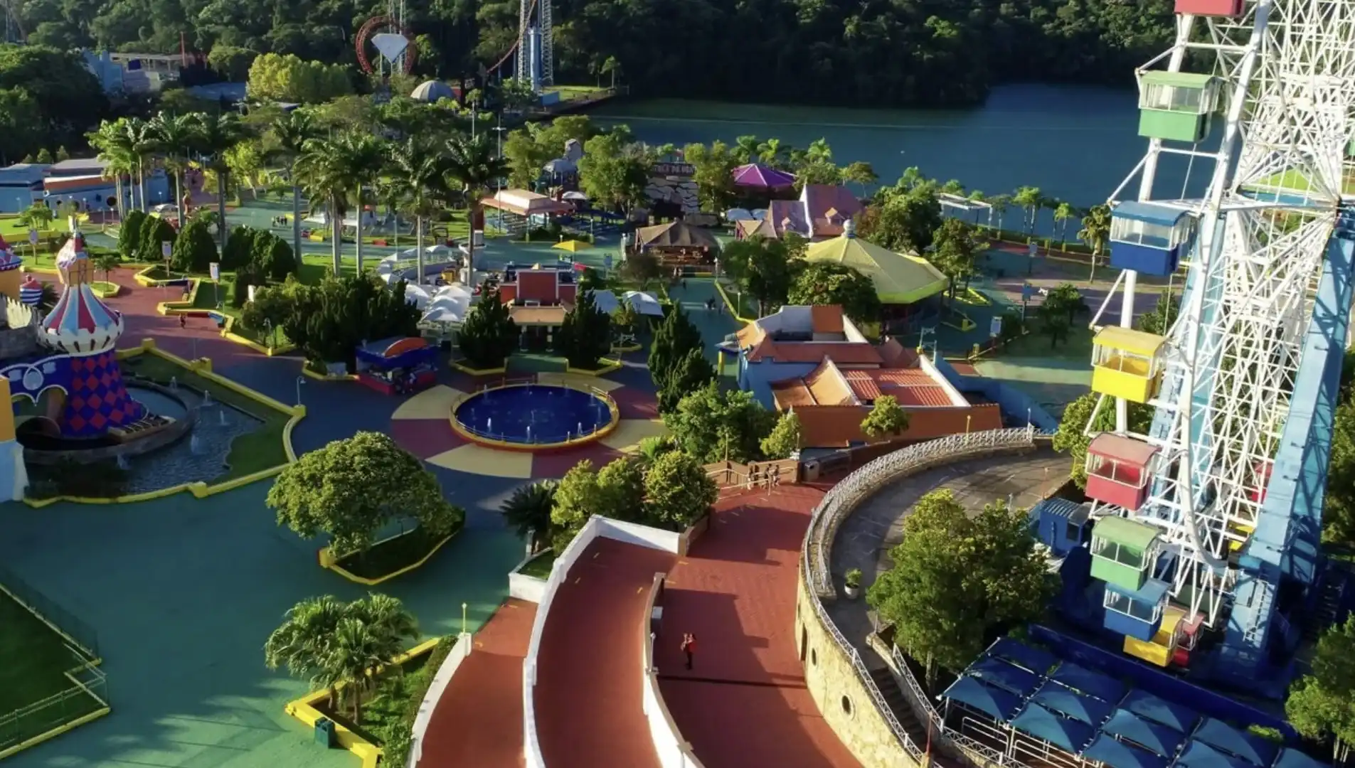 Vista aérea panorâmica do parque de diversões Hopi Hari, mostrando uma roda-gigante colorida, caminhos arborizados, fontes e um lago ao fundo.