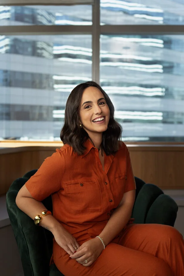 Uma mulher hispânica de meia-idade, com cabelos castanhos ondulados na altura dos ombros e um macacão laranja, sorri alegremente e olha para a direita enquanto está sentada em uma cadeira de veludo verde escuro. Ela está apoiando o cotovelo esquerdo no braço da cadeira e tem a mão direita apoiada no colo. Seu braço esquerdo tem uma pulseira de ouro e seu braço direito tem uma pulseira de ouro e uma pulseira de contas de prata. Ela também tem dois anéis de ouro na mão direita. O fundo é uma janela grande com vista para a cidade. A luz solar brilha na janela e ilumina o quarto.