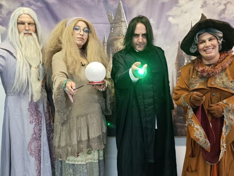 Quatro adultos vestidos como personagens de Harry Potter posam em frente a um pano de fundo do castelo de Hogwarts. O grupo inclui Dumbledore, Trelawney com uma bola de cristal, Snape com uma varinha iluminada e uma quarta pessoa com um chapéu de ponta.