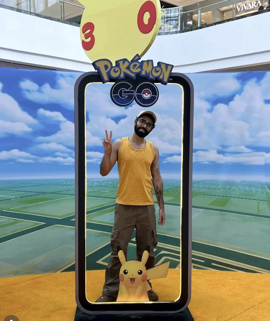 Um homem sorrindo e fazendo o sinal da paz está posicionado dentro de um grande "photo-op" no formato de uma tela de celular gigante. A estrutura tem o logo "Pokémon GO" no topo e a cabeça do Pikachu com os números "3" e "0" acima dela. O fundo dentro do photo-op é um cenário de nuvens e o mapa do jogo. Um pequeno boneco de Pikachu está na base. O homem veste uma regata amarela, calças cargo marrons e um boné. A cena se passa no átrio de um shopping.