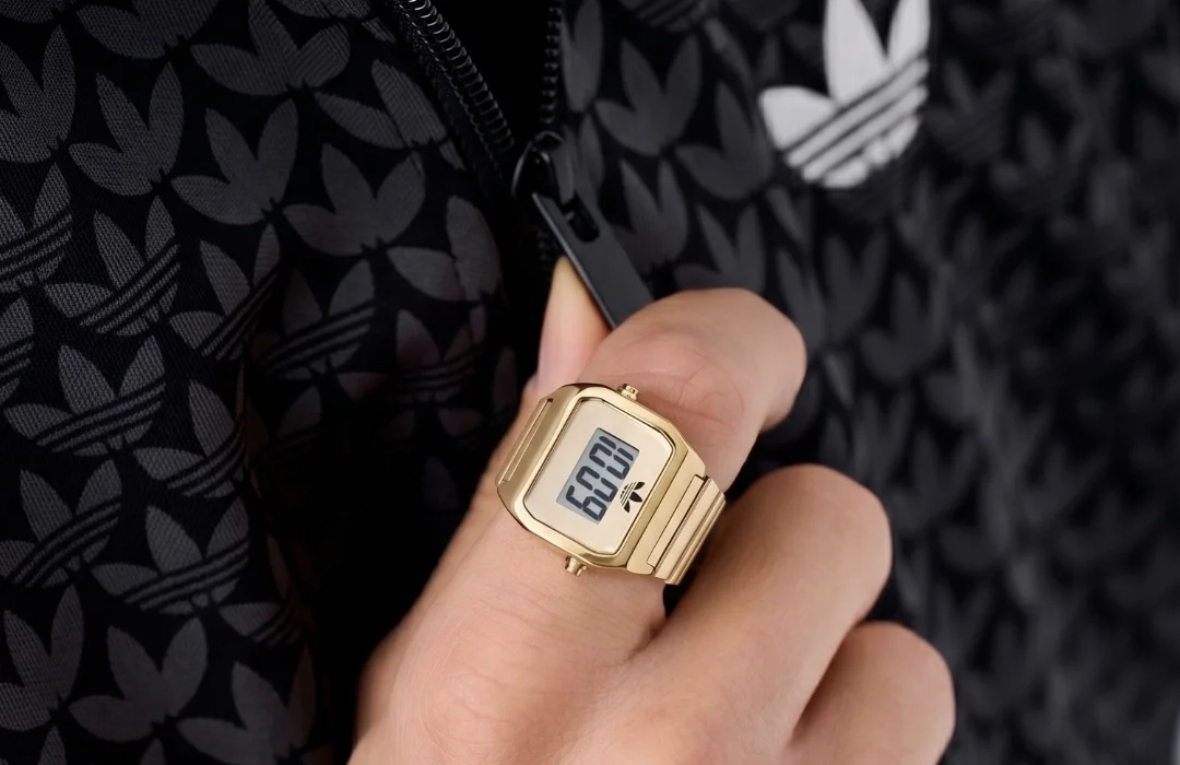 Um close do anel de relógio dourado da Adidas em um dedo, segurando o zíper de uma jaqueta preta com um padrão de monograma da Adidas.