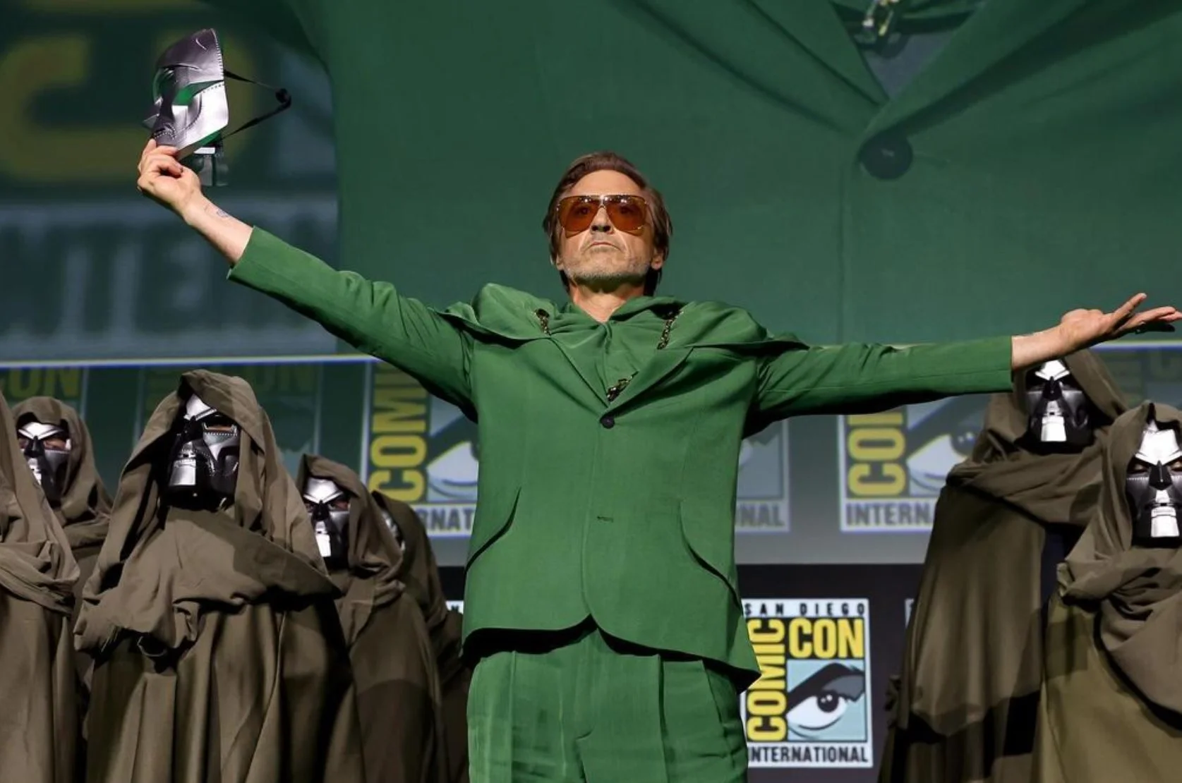 Robert Downey Jr. no palco da San Diego Comic-Con, vestindo um terno verde e erguendo a máscara do Doutor Destino para revelar seu retorno à Marvel.