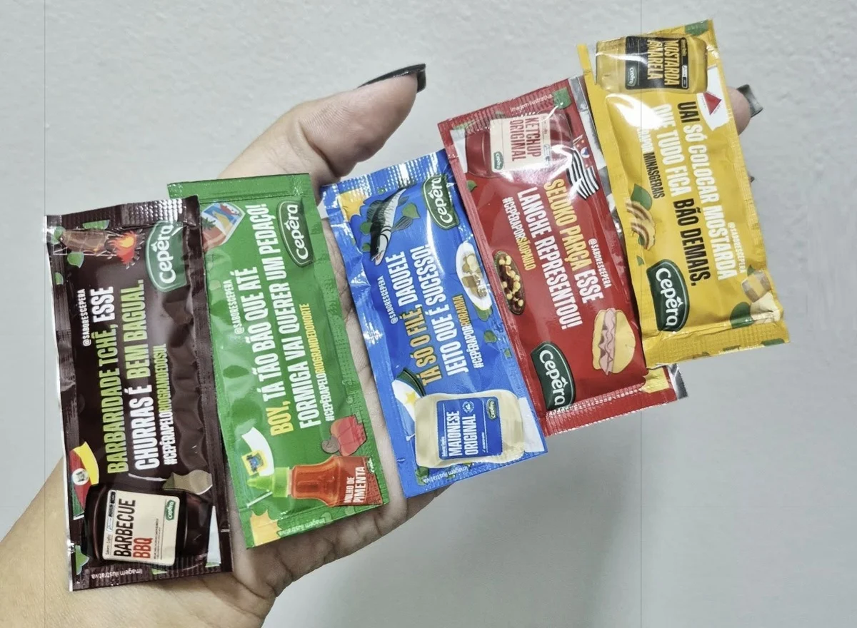 Mão segurando cinco sachês de condimentos da marca Cepêra, cada um com uma cor diferente e frases curtas utilizando gírias regionais brasileiras.