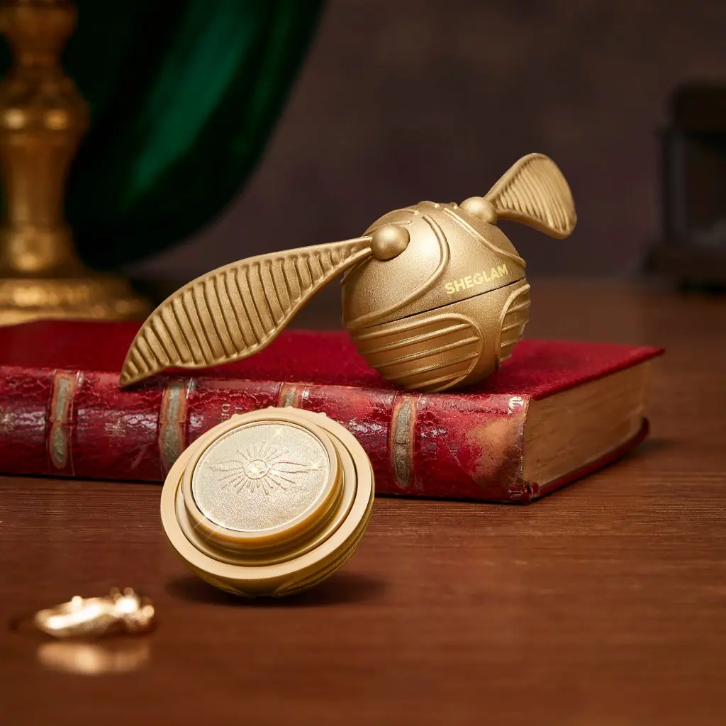 Iluminador dourado em formato de Pomo de Ouro da SHEGLAM posicionado sobre um livro antigo de capa vermelha.