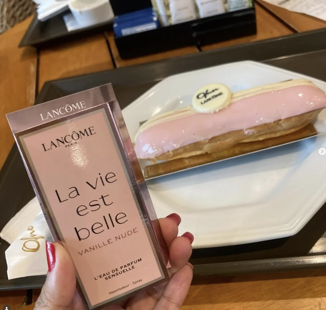 Mão segurando uma amostra do perfume Lancôme "La Vie Est Belle Vanille Nude" em frente a um prato com um éclair decorado em uma bandeja.