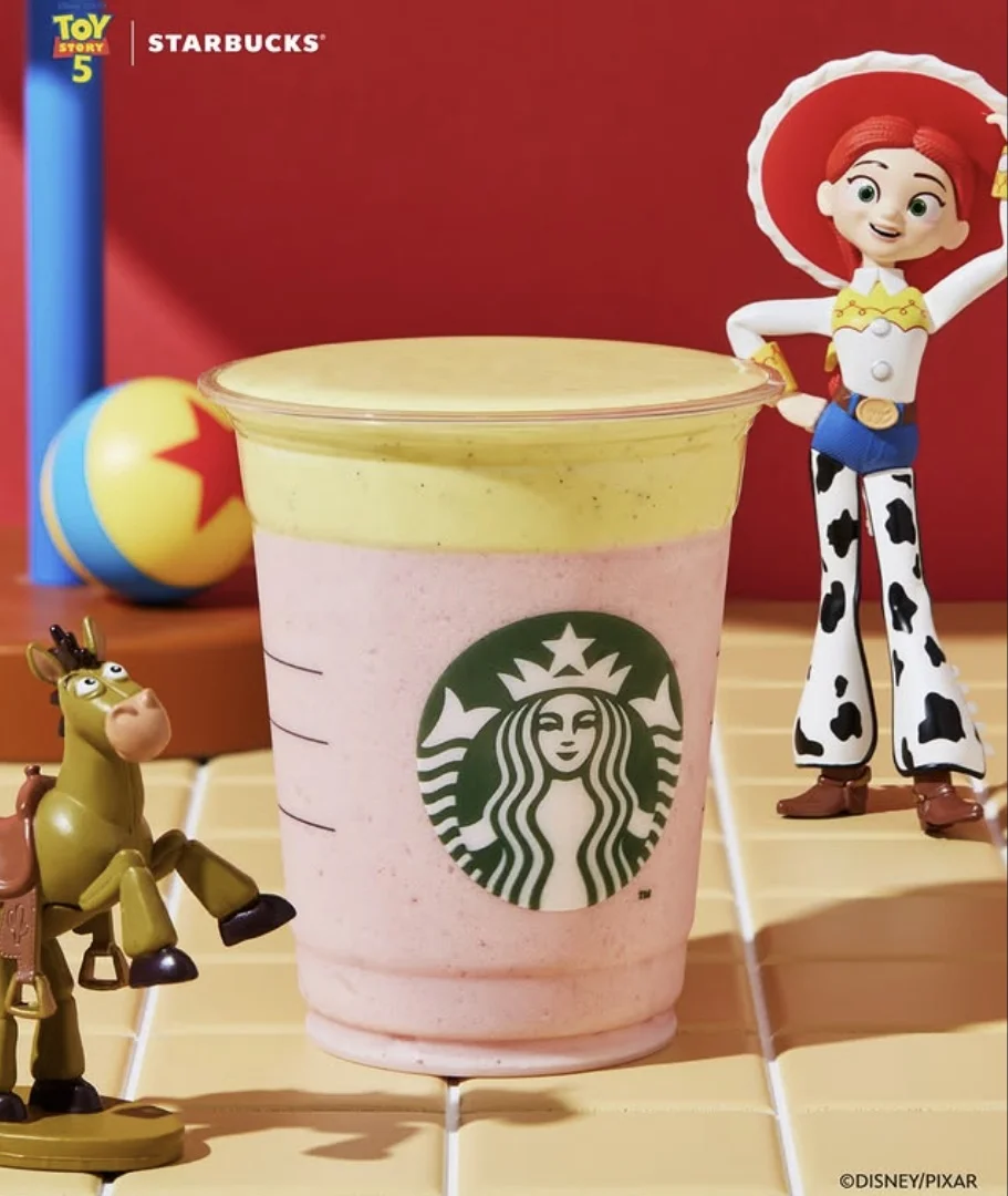Um copo de plástico transparente da Starbucks cheio de uma bebida de duas camadas, rosa e amarela, cercado por brinquedos de Toy Story e um logotipo da colaboração.