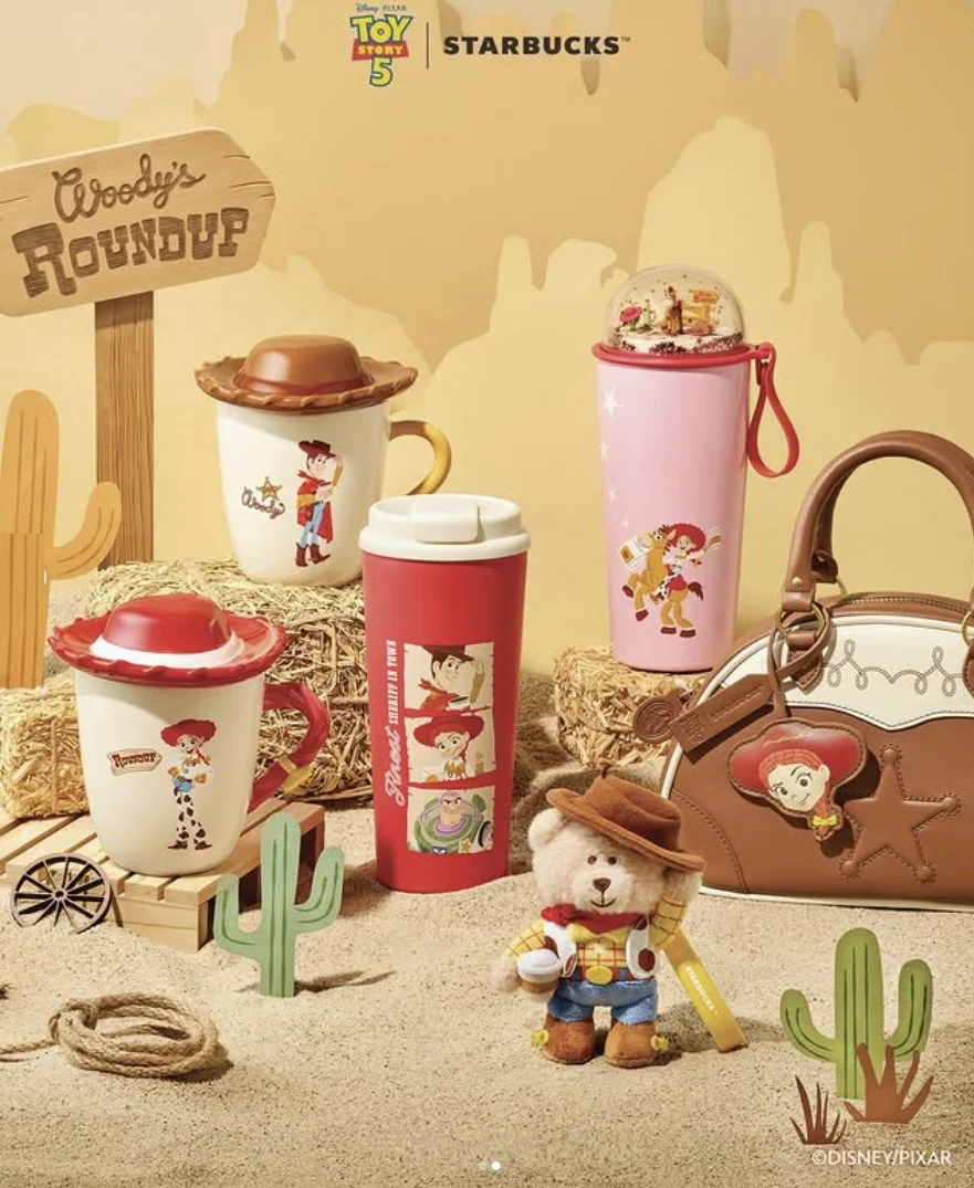 Uma coleção de produtos Starbucks x Toy Story com tema de "Woody's Roundup", incluindo canecas, garrafas e uma bolsa, dispostos em um cenário de faroeste.