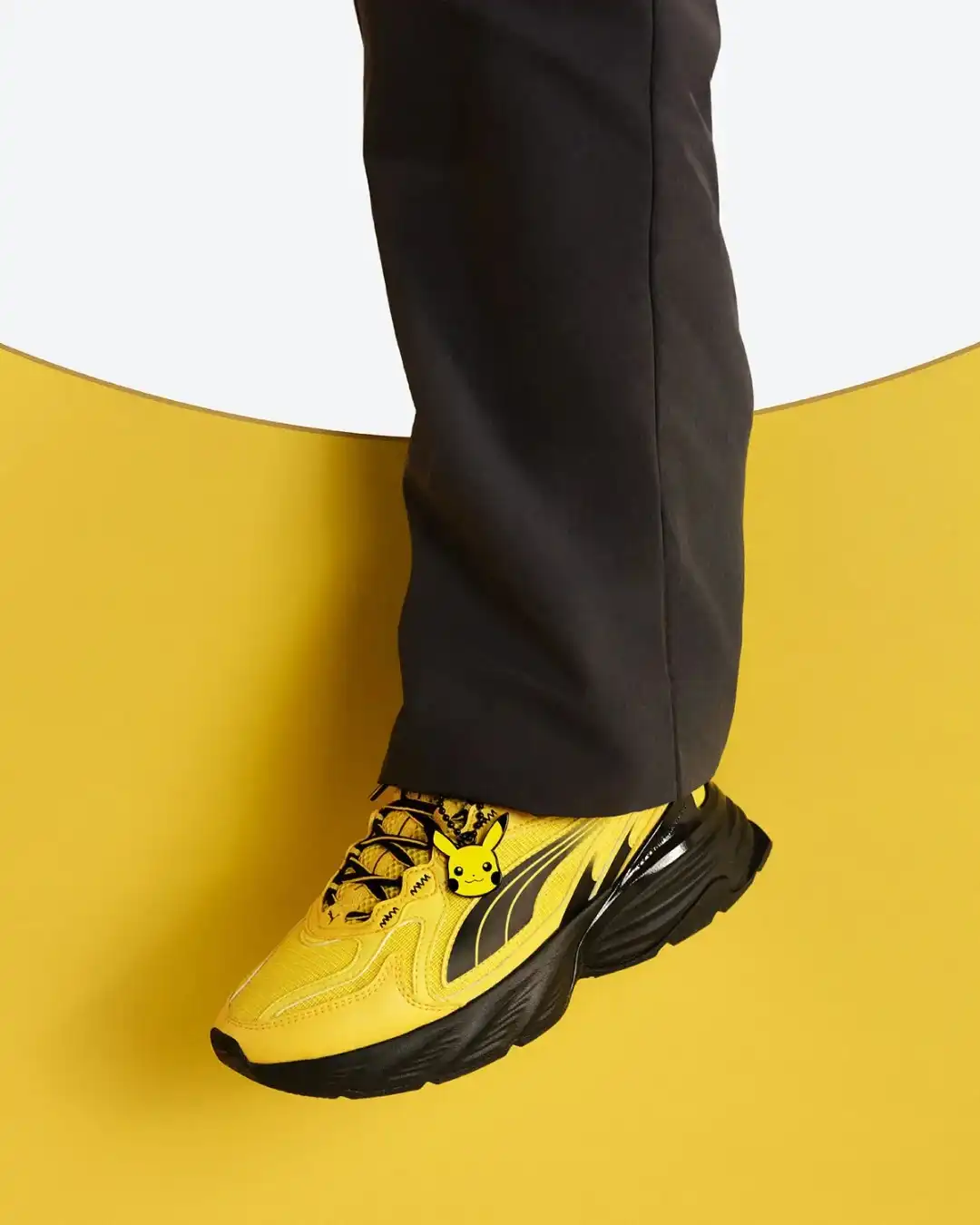 Pessoa vestindo calça preta calçando o tênis Puma amarelo do Pikachu contra um fundo circular amarelo.