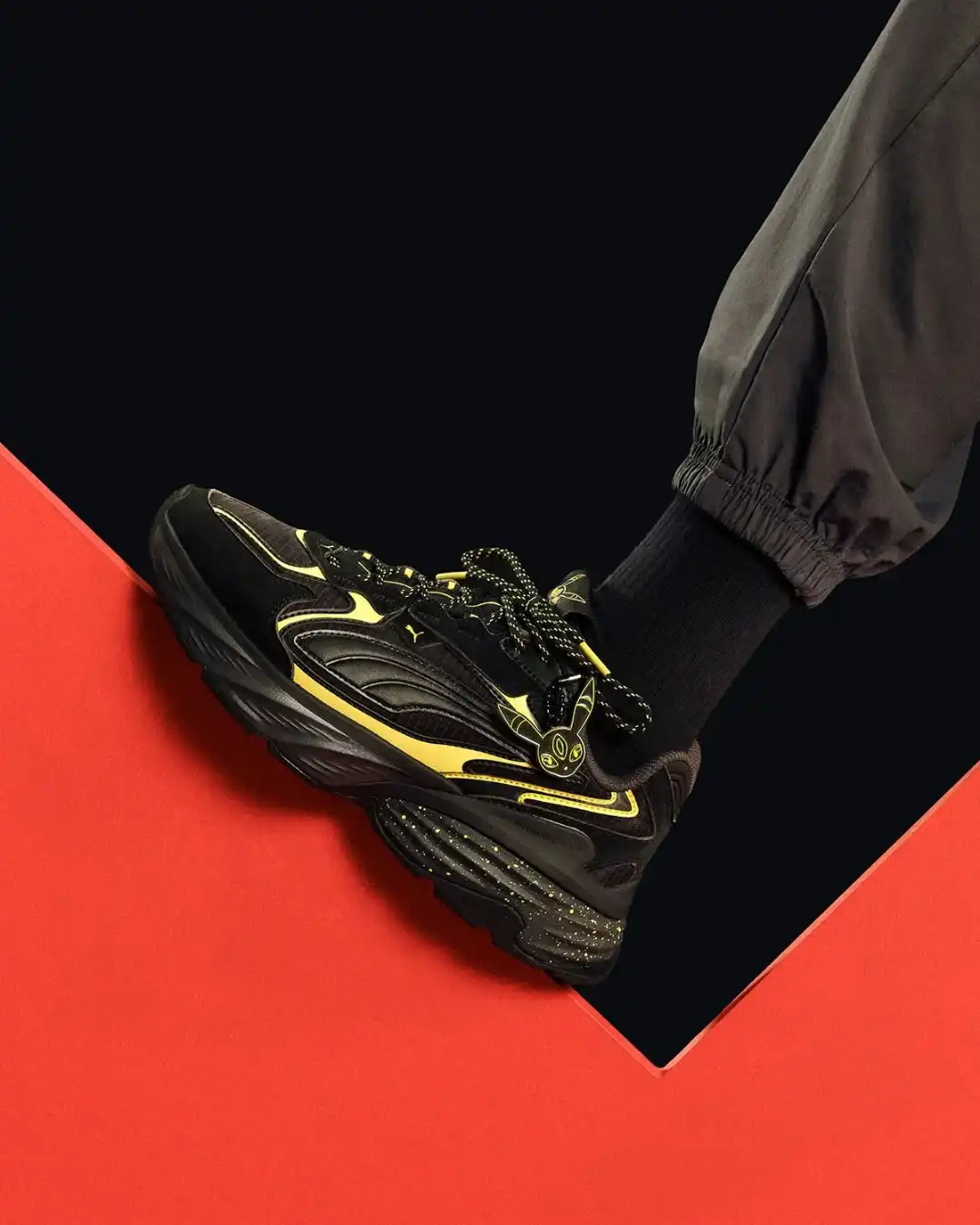 Tênis Puma preto com detalhes amarelos e respingos na sola, sendo usado sobre uma superfície geométrica vermelha e preta.