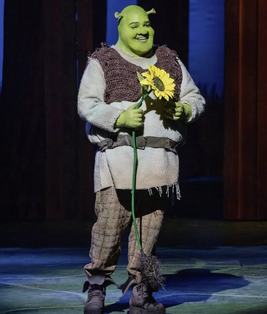 Uma imagem de um ator vestido como Shrek, em pé em um palco de teatro e segurando um girassol. O ator está usando um traje completo de Shrek, incluindo maquiagem verde, orelhas de ogro e roupas rústicas. Ele está sorrindo e olhando para o girassol.