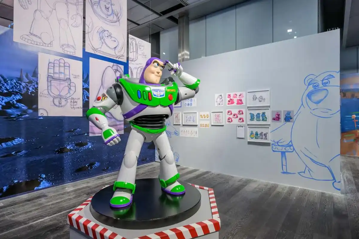 Uma estátua em tamanho real do personagem Buzz Lightyear, de Toy Story, em uma exposição da Pixar, fazendo sua pose de saudação característica com um dedo na têmpora. Ele está em pé sobre um plinto octogonal com bordas vermelhas e brancas. O fundo apresenta arte conceitual e uma grande silhueta do Urso Lotso.