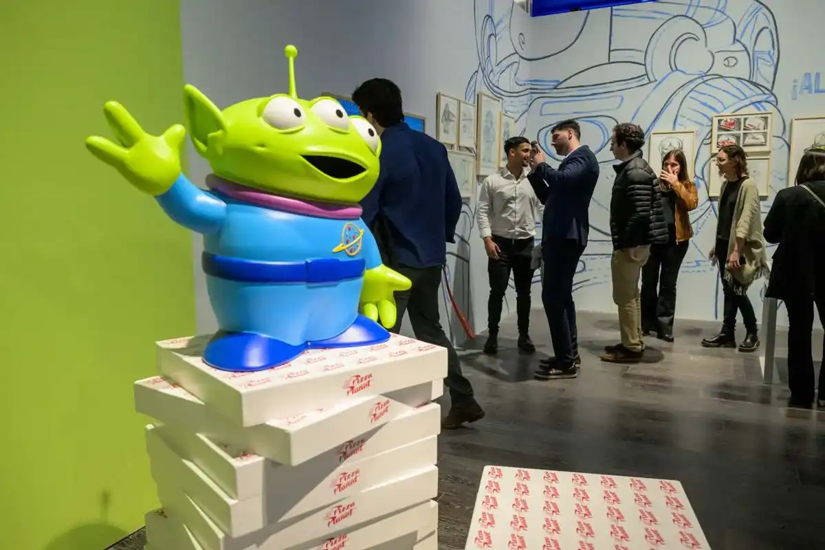 Uma estátua grande de um Alienígena de Três Olhos, de Toy Story, em pé sobre uma pilha de caixas de pizza Pizza Planet em uma exposição da Pixar. O alienígena está acenando com uma mão e tem um sorriso aberto. Pessoas estão de pé ao fundo, observando a arte conceitual nas paredes.