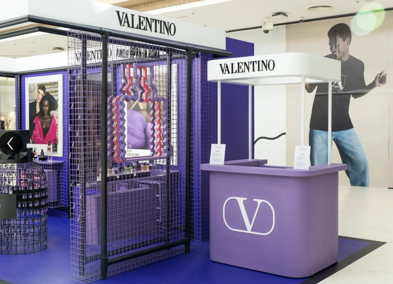 Uma vista angular do estande da Valentino no shopping. À esquerda, uma das instalações de garrafas de perfume exageradas feitas de cadeados lilases. No centro-esquerda, um display de malha de arame roxa contém prateleiras com várias garrafas de perfume e uma instalação geométrica em V-Studs rosa e roxo. À direita, um quiosque de servir separado e modular, de cor lilás suave, com o logotipo "V" branco da Valentino e a inscrição "VALENTINO" no dossel superior branco. Dois panfletos de menu estão no balcão. Um grande pôster borrado de um homem de camisa preta está em segundo plano. O chão é o carpete azul cobalto.