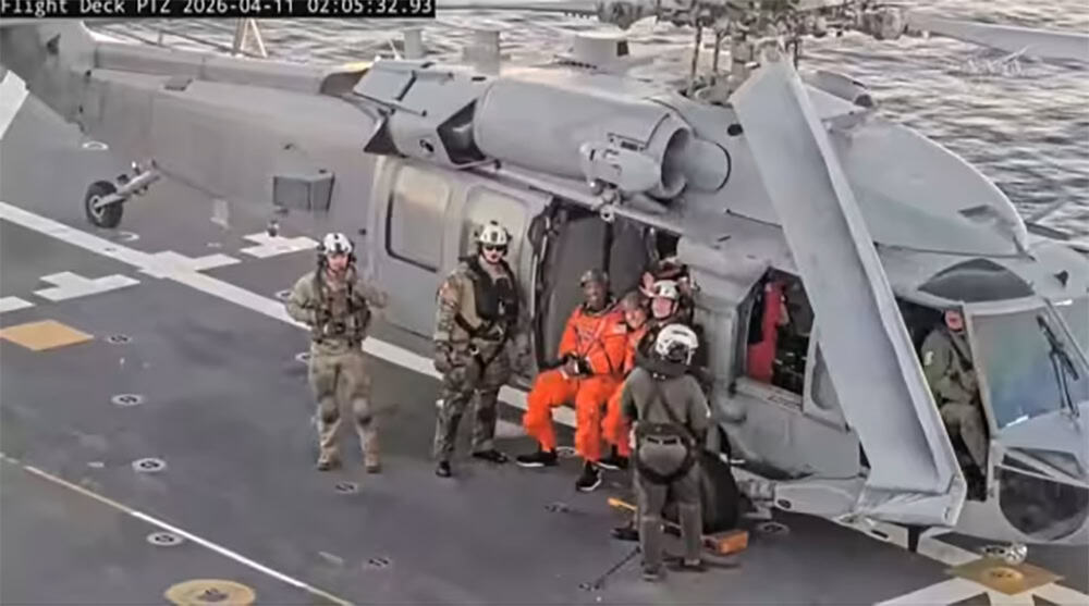 Uma imagem de satélite aproximada de quatro astronautas com roupas de voo laranja brilhante saindo da espaçonave Orion e subindo em um bote salva-vidas hexagonal inflável laranja e preto no Oceano Pacífico.