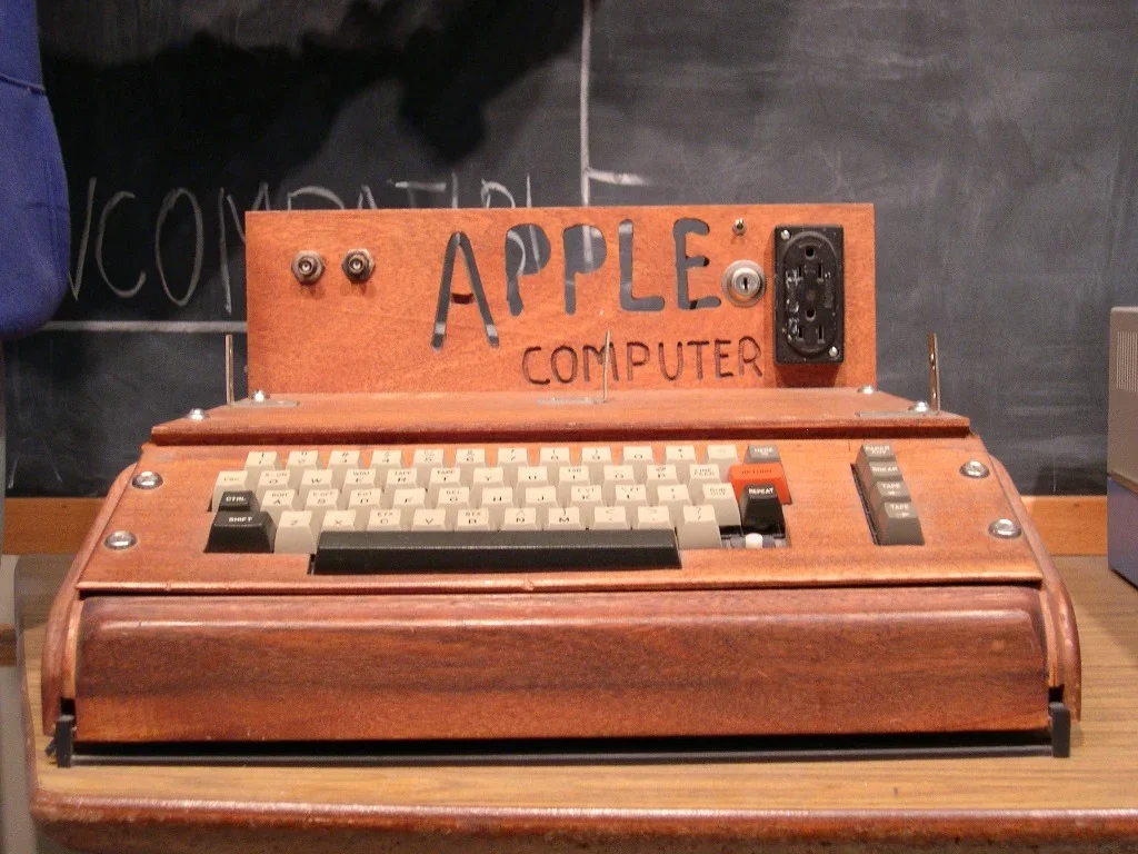 Uma foto em close de um computador Apple I original, alojado em uma caixa de madeira artesanal com um teclado QWERTY completo e uma placa de identificação com o texto "APPLE COMPUTER" gravado. Ele está sobre uma mesa de madeira em um laboratório ou oficina.