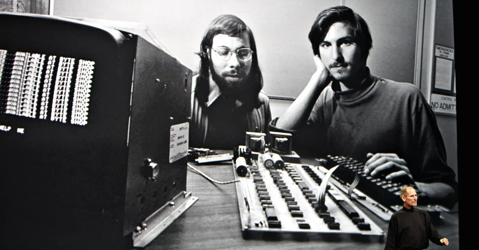 Uma foto em preto e branco de Steve Jobs e Steve Wozniak em meados da década de 1970, sentados em um escritório com um computador de tela grande, um Apple I e outros componentes eletrônicos. No canto inferior direito, há uma pequena imagem colorida de Steve Jobs no palco.