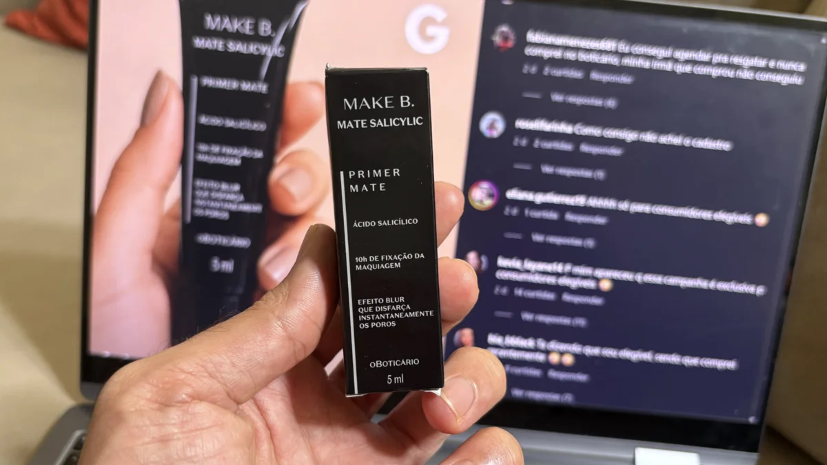 Uma mão segura uma pequena embalagem preta do primer 'Make B. Mate Salicylic' em frente a um notebook que exibe comentários de uma rede social sobre a campanha de brindes do Boticário