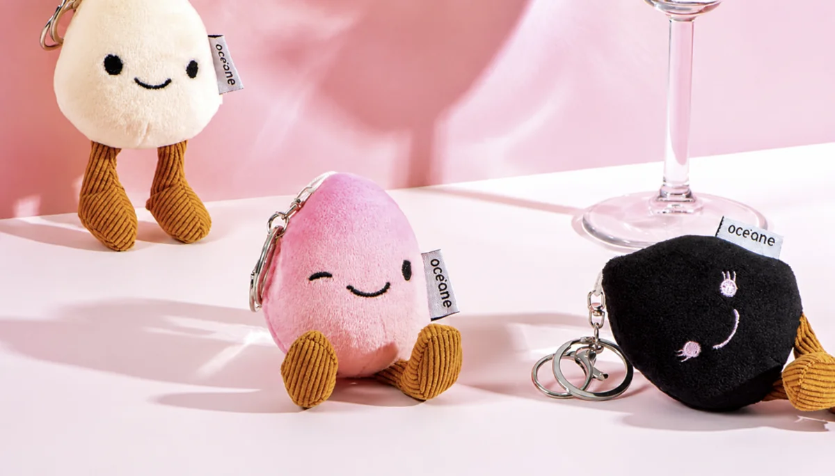 Três chaveiros de pelúcia em formato de esponja (nas cores bege, rosa e preto) sentados em uma superfície branca sob luz rosada. Eles possuem rostos bordados e etiquetas com a marca "Océane".
