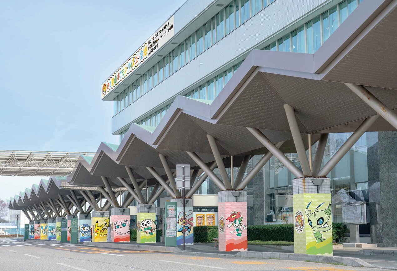 Fachada externa do Aeroporto Noto Satoyama, no Japão, decorada com colunas coloridas ilustrando vários personagens de Pokémon, sob uma cobertura ondulada cinza.