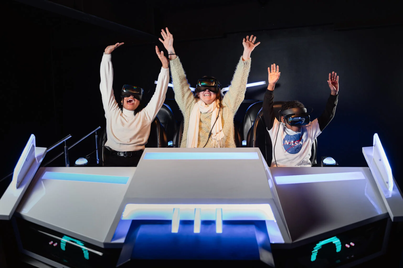 Três pessoas sentadas em um simulador de movimento, usando óculos de realidade virtual e com os braços erguidos em sinal de comemoração e euforia.