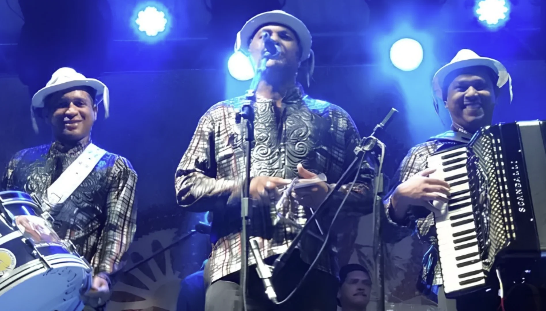 Um trio de músicos de forró se apresenta em um palco com iluminação azul.