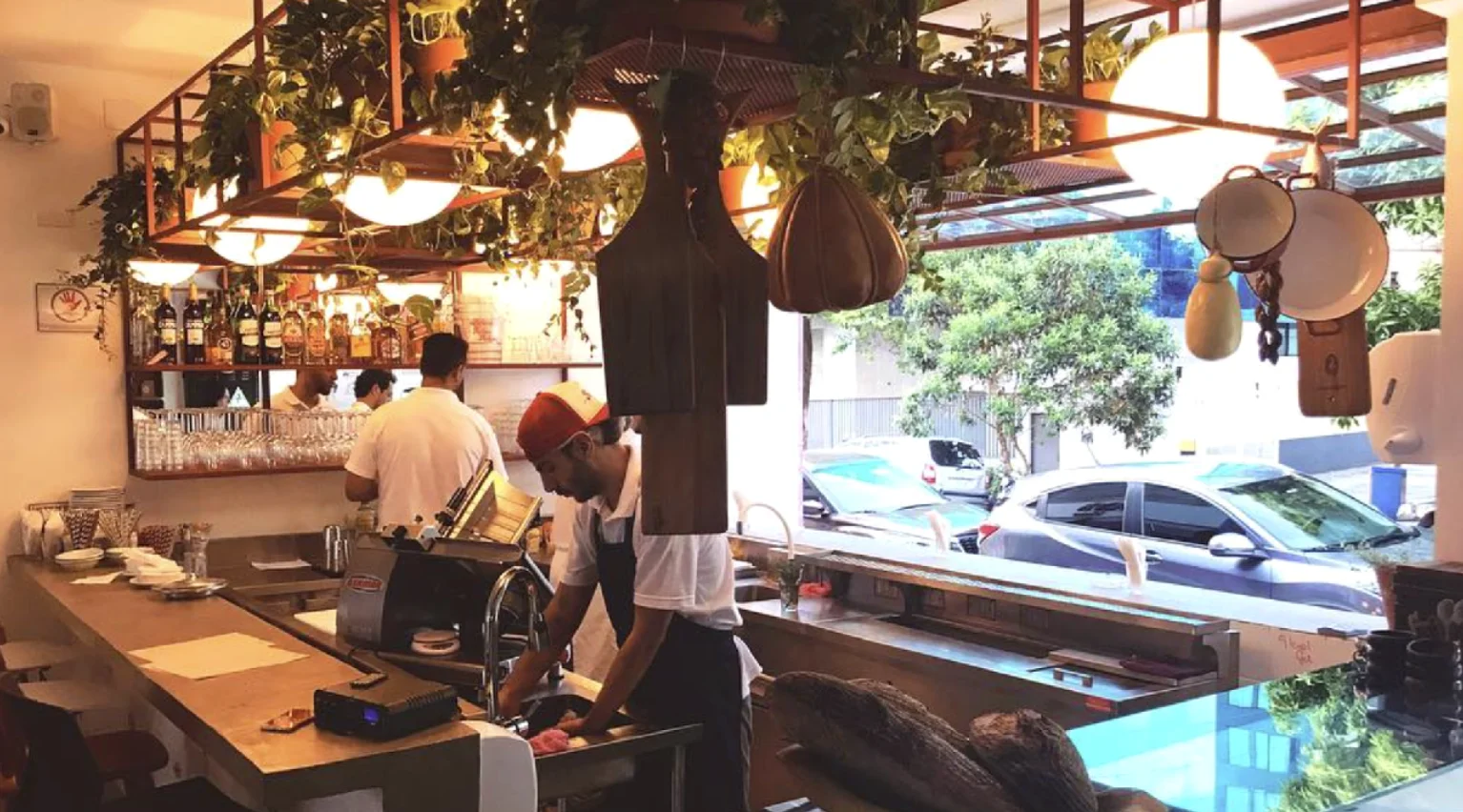 Interior iluminado de um bar e mercearia moderno, mostrando funcionários trabalhando atrás de um balcão de madeira comprido e plantas pendendo do teto.