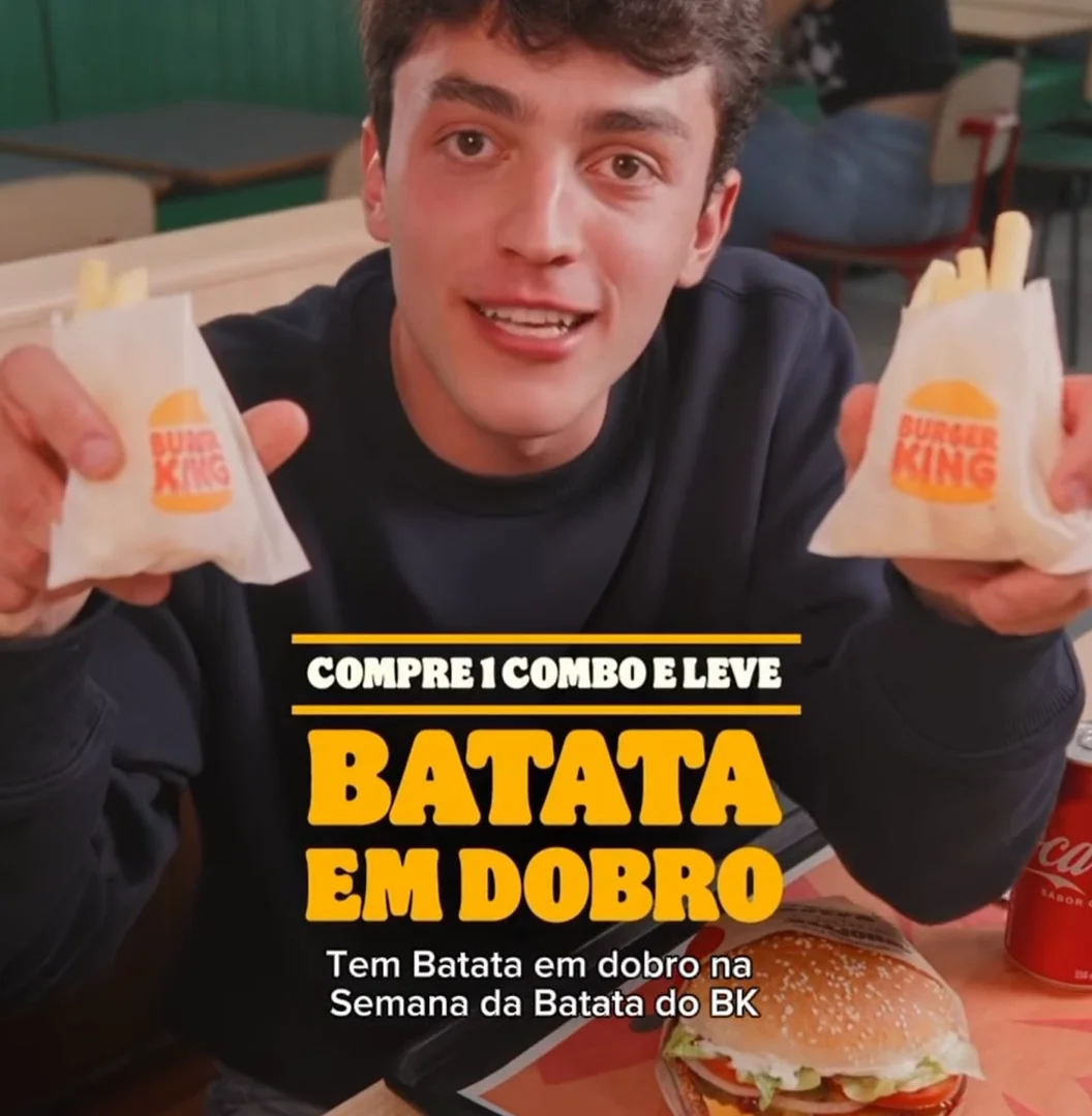 Homem jovem sorrindo em um restaurante Burger King, segurando duas embalagens de batata frita, com um hambúrguer e uma lata de Coca-Cola na mesa à sua frente.