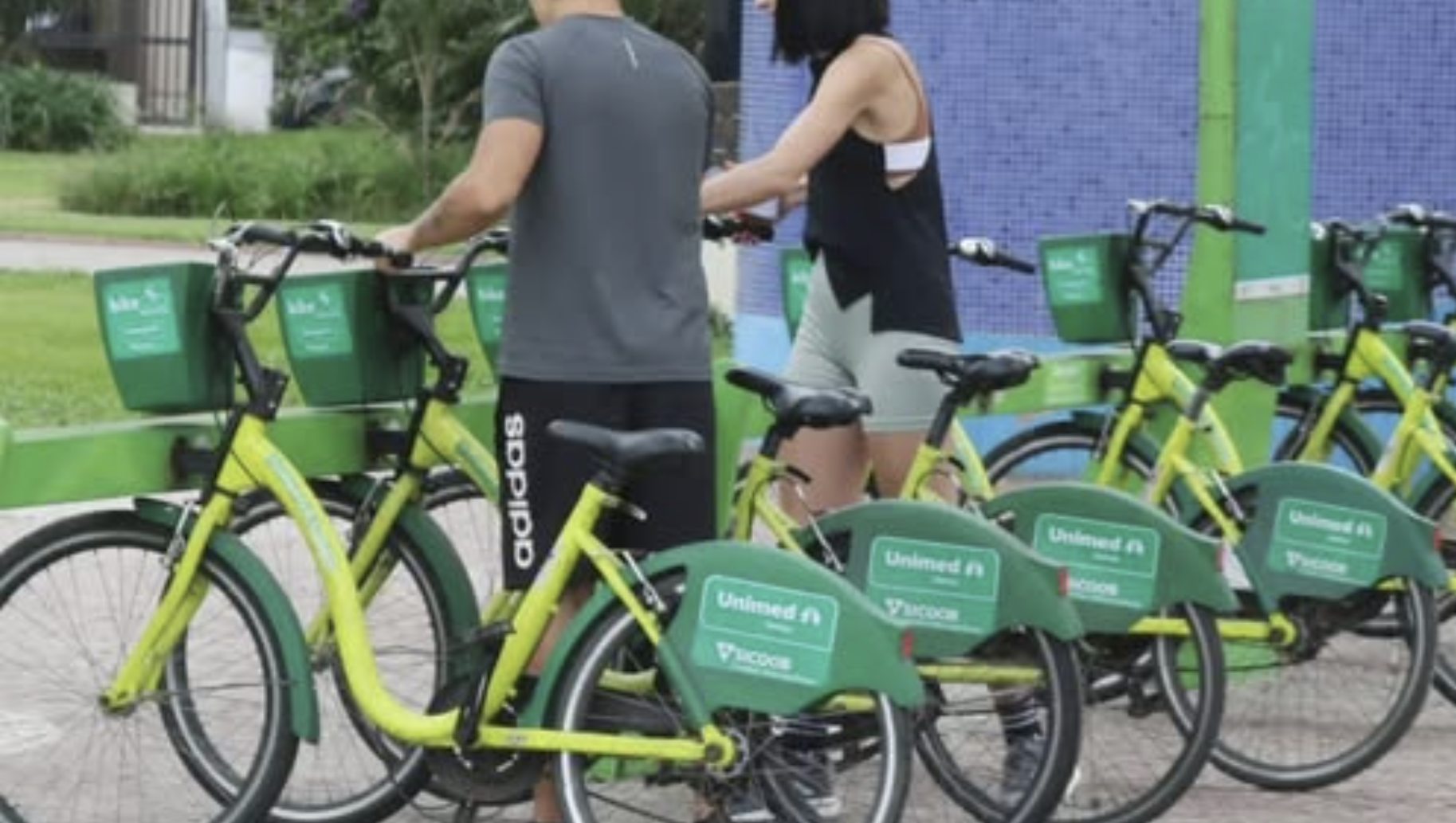 Duas pessoas de costas retirando bicicletas compartilhadas de cor verde limão e verde escuro em uma estação de autoatendimento ao ar livre.