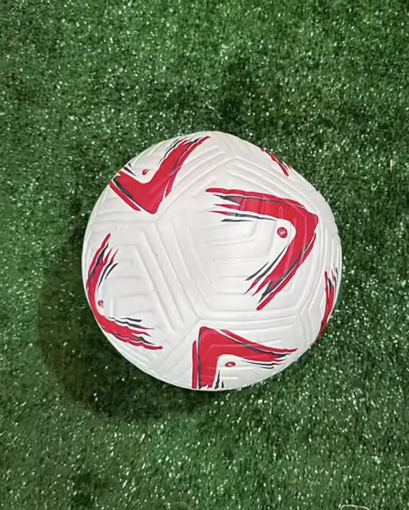 Bola de futebol branca com detalhes em vermelho e cinza, posicionada sobre um gramado sintético verde, vista de cima.
